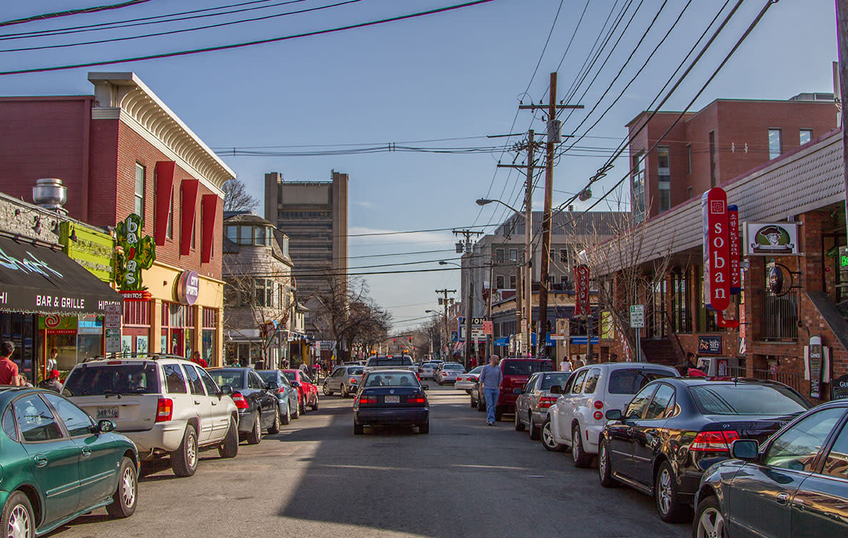 Street view of Thayer Street in Providence, Rhode Island, showing parked cars, storefronts, apartment buildings, street signs, utility lines, and steady traffic under a clear daytime sky.