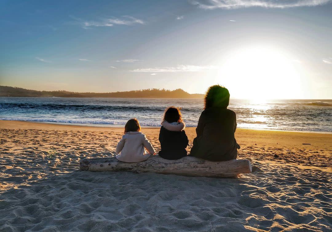 Mother and her two daughters sitting on a log facing the beach while the sun sets.