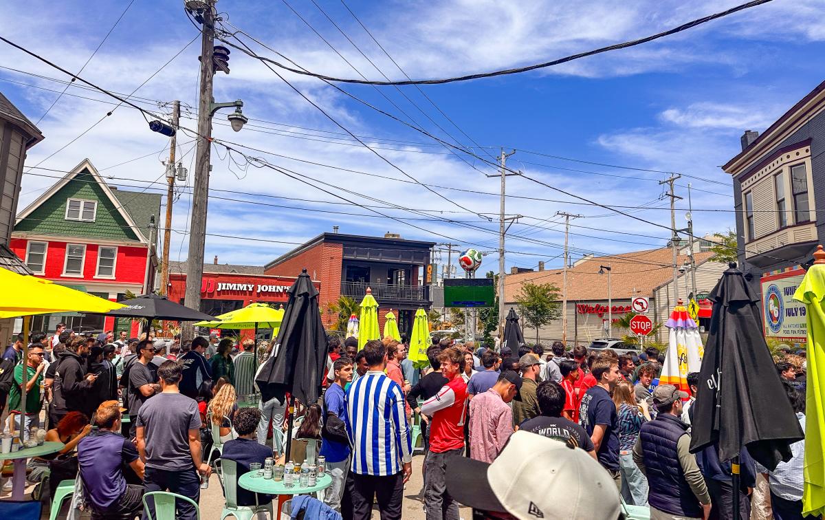 Soccer fans pack the patio and surrounding area outside Nomad World Pub on Brady Street in Milwaukee, watching a match on a large outdoor screen during a sunny afternoon.