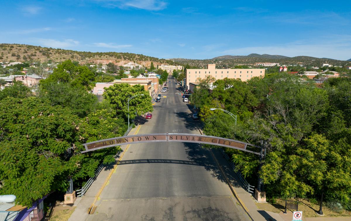 A view of a street in Silver City, New Mexico, lined with trees on both sides. A sign arches over the road reading "Downtown Silver City." In the distance, hills and several low-rise buildings are visible under a clear blue sky.