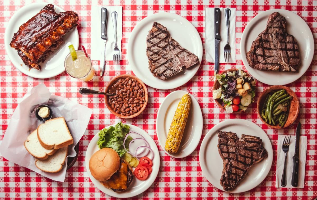 top view of a table with plates of steak, ribs, corn on the cob, beans, burgers, bread, and butter