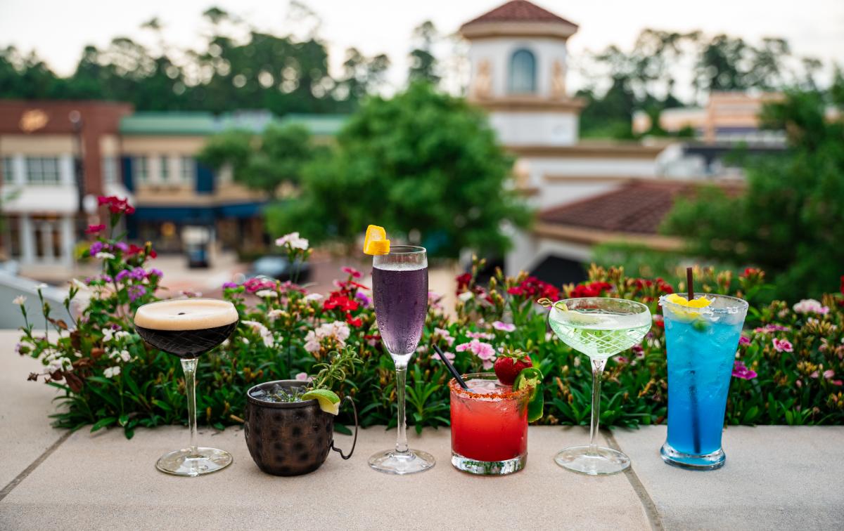 Six colorful drinks are lined up on the balcony at Hyatt Centric's rooftop pool. On the far left, an espresso martini, then a mule with lime and sprig of rosemary, then a tall purple cocktail, then a bright red drink with a strawberry and jalapeno slice on the rim, then a cucumber martini, then a tall blue drink. In the background is an (out of focus) overview of Market Street.