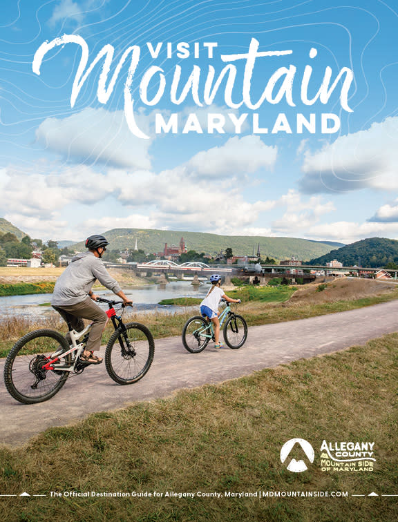 Cover of the Visit Mountain Maryland Destination Guide showing an adult and child biking along the C&O Canal Towpath in Cumberland, Maryland, with a river, bridge, downtown, and mountains in the background, and the text “Visit Mountain Maryland.”
