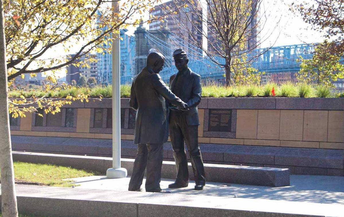 Statue of two men in Union Civil War uniforms reaching for each other with the Roebling Suspension Bridge in the background