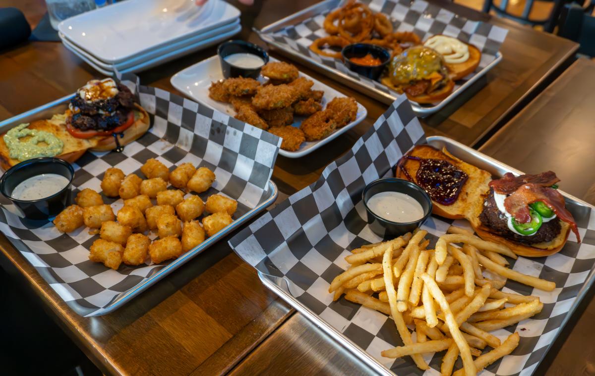 A table filled with diner-style comfort food, including burgers topped with bacon and cheese, crispy chicken wings with dipping sauce, golden tater tots, onion rings, and a side of French fries, all served on trays lined with checkered paper.