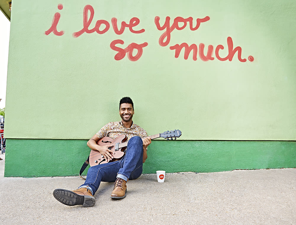 Musician Tje Austin sits below a mural reading "I love you so much." on South Congress Ave. He is holding a guitar and has a cup of coffee sitting next to him