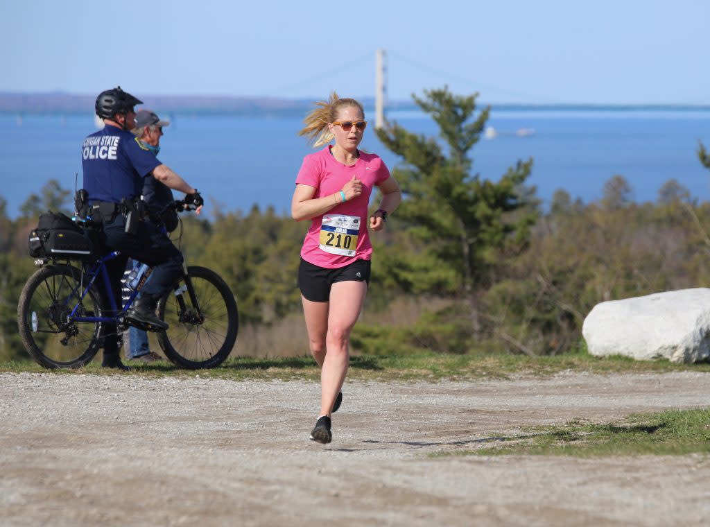 A woman runs around a corner and uphill during a Mackinac Island road race with the water and Mackinac Bridge in the background