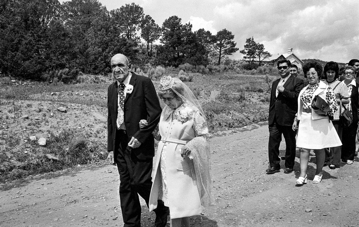 Black and white photograph of an elderly couple dressed in formal clothes walking arm in arm.