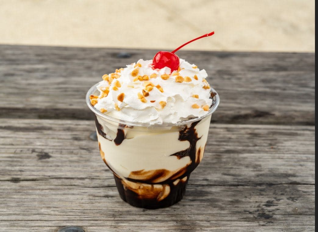 A hot fudge sundae from Moosa's Frozen Custard sits on a weathered wooden table, layered with vanilla frozen custard and chocolate sauce, then topped with whipped cream, chopped peanuts and a maraschino cherry.