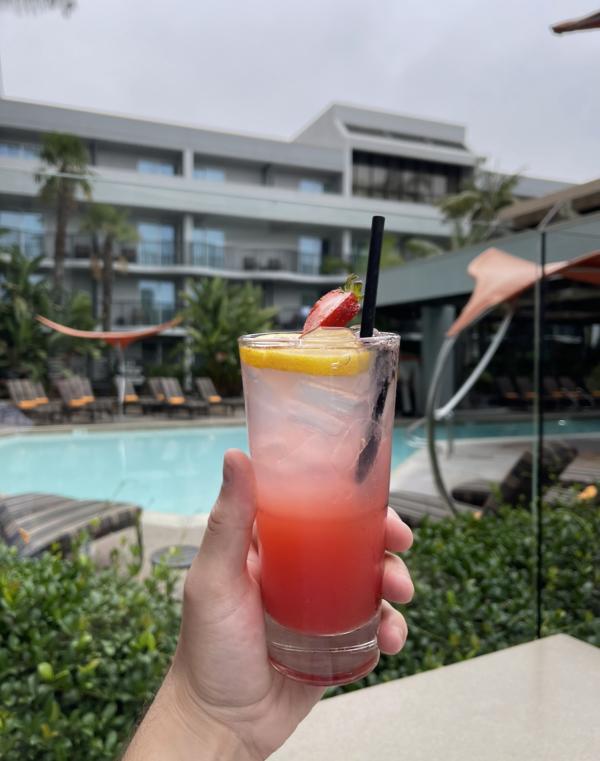 Image of a red-ish and pink-ish cocktail being held up. In the background, a pool and pool chairs can be seen.