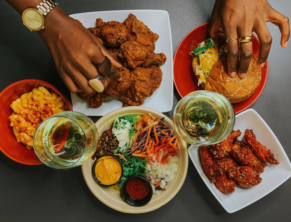 Black woman's hands reaching for a plate of crispy fried chicken and loaded burger. White wine glasses, mac and cheese, chicken wings and a Korean barbecue bowl are also on the table.