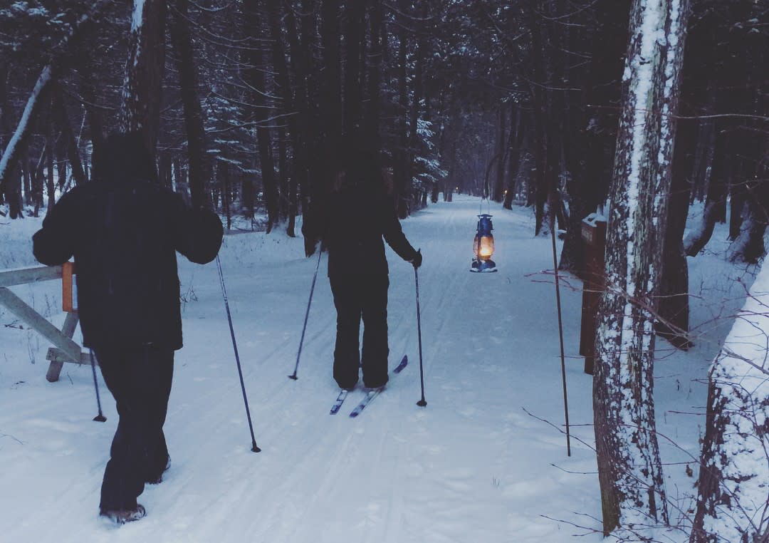 Two cross-country skiers pass by a lantern in the woods of Mackinac Island State Park during the Twilight Turtle Trek
