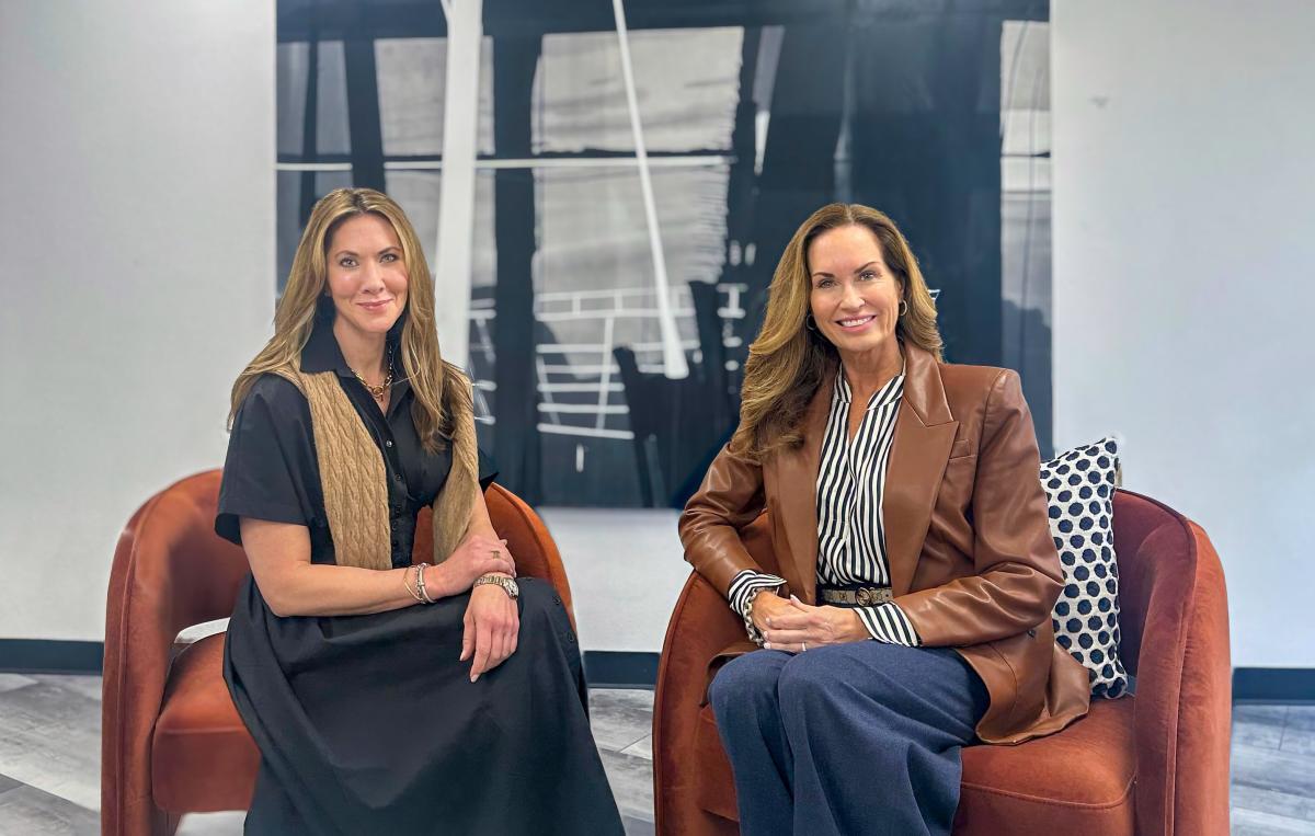 Two women, 30s-40s, smile at the camera. They both have long dirty-blonde hair and are sitting on burned-orange cushioned chairs before a black-and-white abstract art piece. The woman on the left is wearing a long, simple black dress with a tan sweater vest. The woman on the right is wearing a black-and-white striped button-down blouse, navy pants, and a brown pleather jacket.