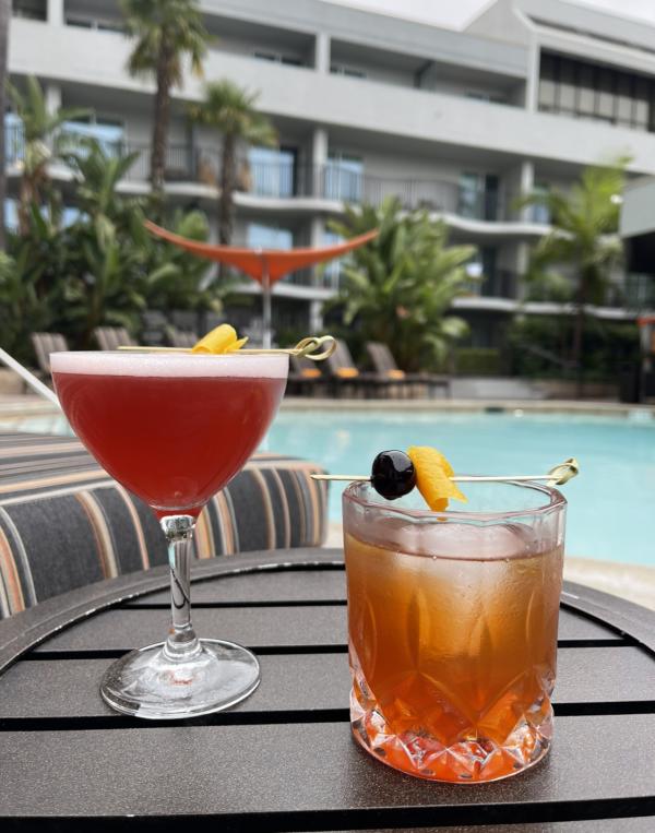 Image of two cocktails placed poolside. The cocktail to the left is purple and served with a lemon peel. The second cocktail, to the right, is a brown-ish and orange-ish color. It is served with an orange peel and dark red cherry.