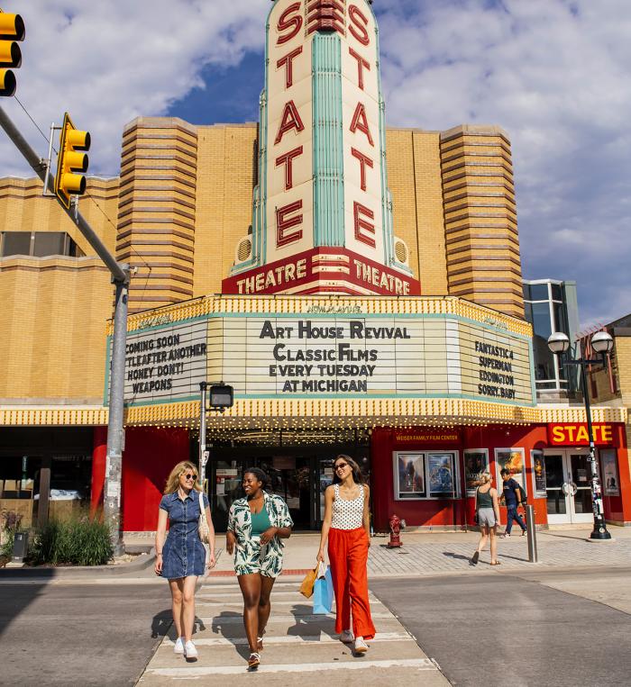 Three women in colorful clothing crossing the crosswalk in front of State Theatre.