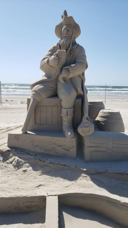 Up close of a sand sculpture of a pirate with a peg leg drinking beer on a pile of wooden debris and treasure chest.