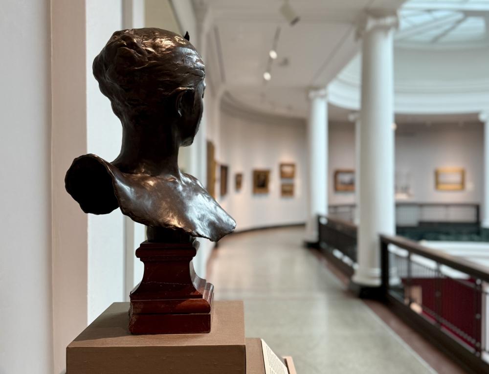 Bronze bust of woman's upper body faces away from the camera in an art museum, with blurred background and paintings hanging on the curved wall in the distance.