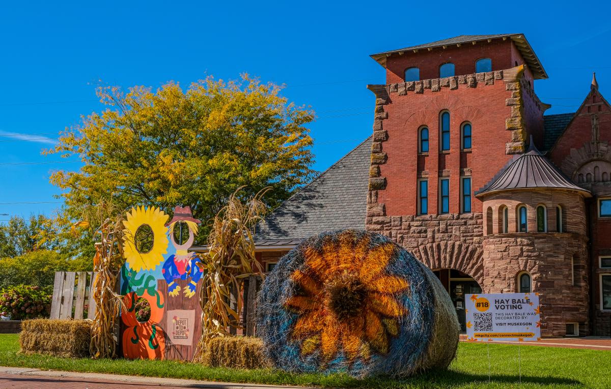 Historic red brick building in richardsonian romanesque style architecture sits behind fall decor of scarecrown cutouts and sunflower made of haybale