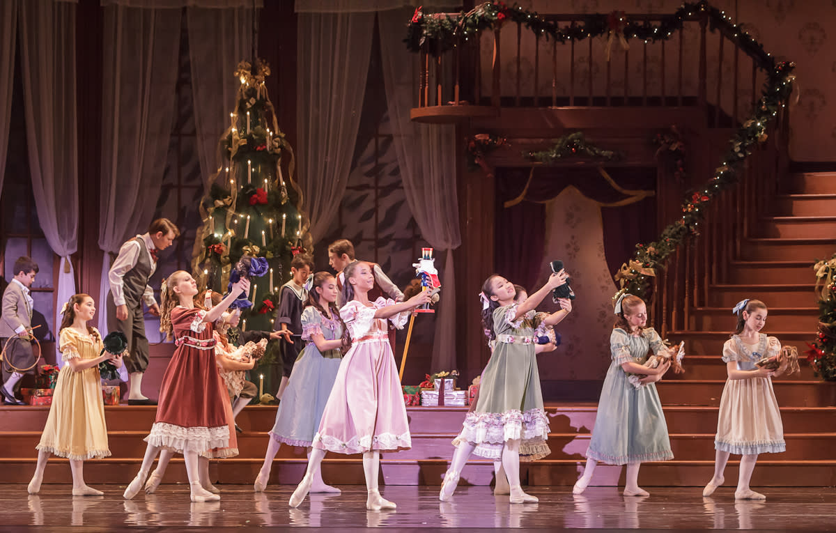 Young girls on stage holding dolls while young boys look at a Christmas tree in the background.