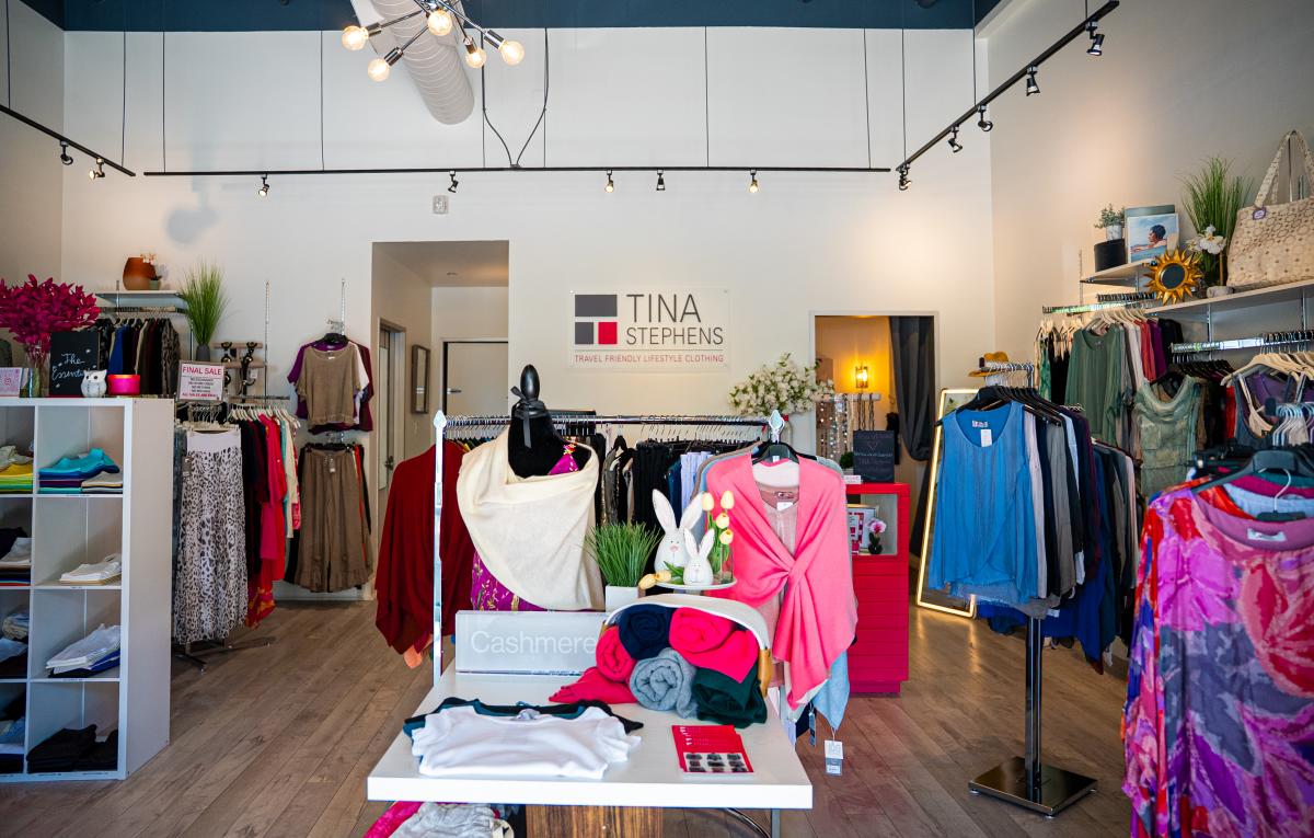 The camera overlooks the interior of the TINA Stephens boutique. It features multiple racks of colorful, stylish, travel-friendly clothes as well as some accessories, bags, mirrors, and other knickknacks. Along the back wall is signage that reads, "TINA STEPHENS | TRAVEL FRIENDLY LIFESTYLE CLOTHING."