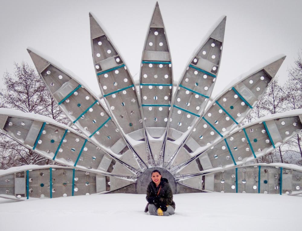 Woman sits in snow in front of sculpture made of nine canoes forming an arch.