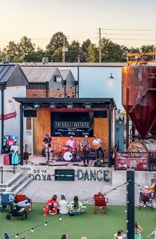 A live band performs on the outdoor stage at Stovehouse as guests relax on the lawn and gather around the venue. The evening scene features string lights, the Gas Light Alley area, and the industrial backdrop of the historic Stovehouse campus.