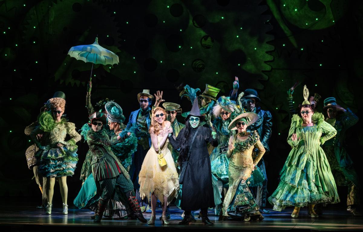 A theatrical scene from the musical Wicked showing the main cast in elaborate green, teal, and yellow costumes on a starlit stage. The ensemble surrounds two central characters - one in a flowing yellow dress and another in dark robes - creating a magical Oz atmosphere with whimsical period costumes.
