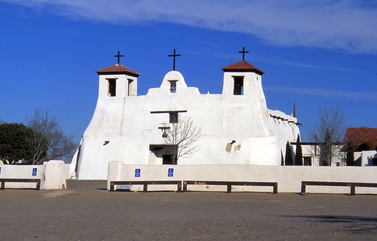 A white adobe church with two steeples topped with crosses sits beneath a blue sky