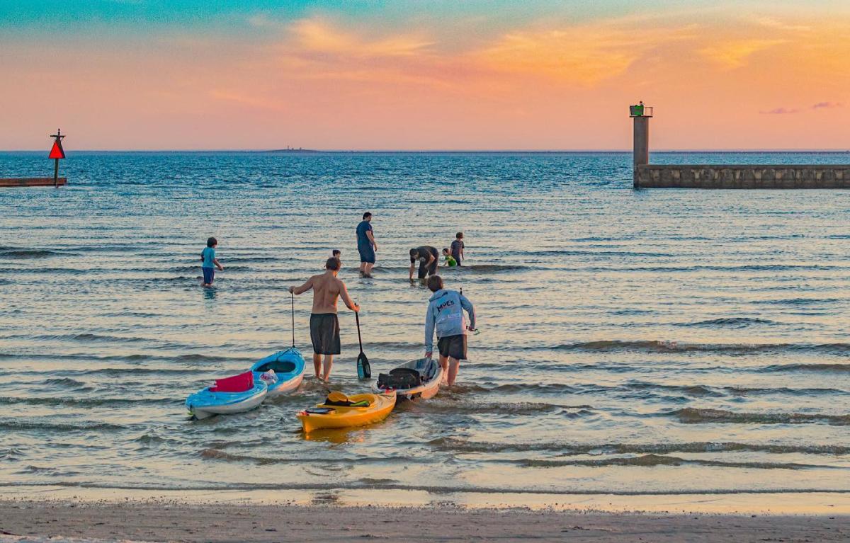 Boys wade into Lake Pontchartrain towing small kayaks behind them. A family wades further out. It is sunset.