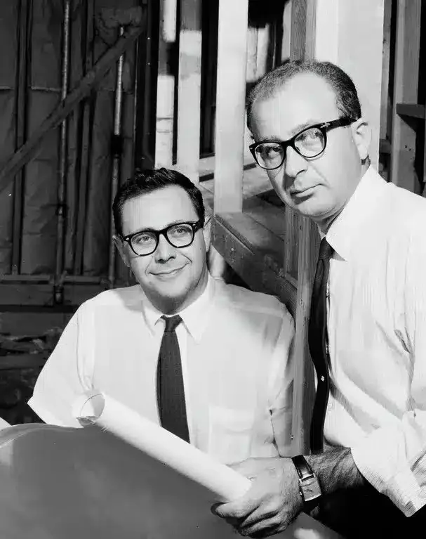 Two men wearing glasses and dress shirts with ties, one holding architectural plans, standing together in a room with wooden framing in the background. The image is in black and white.