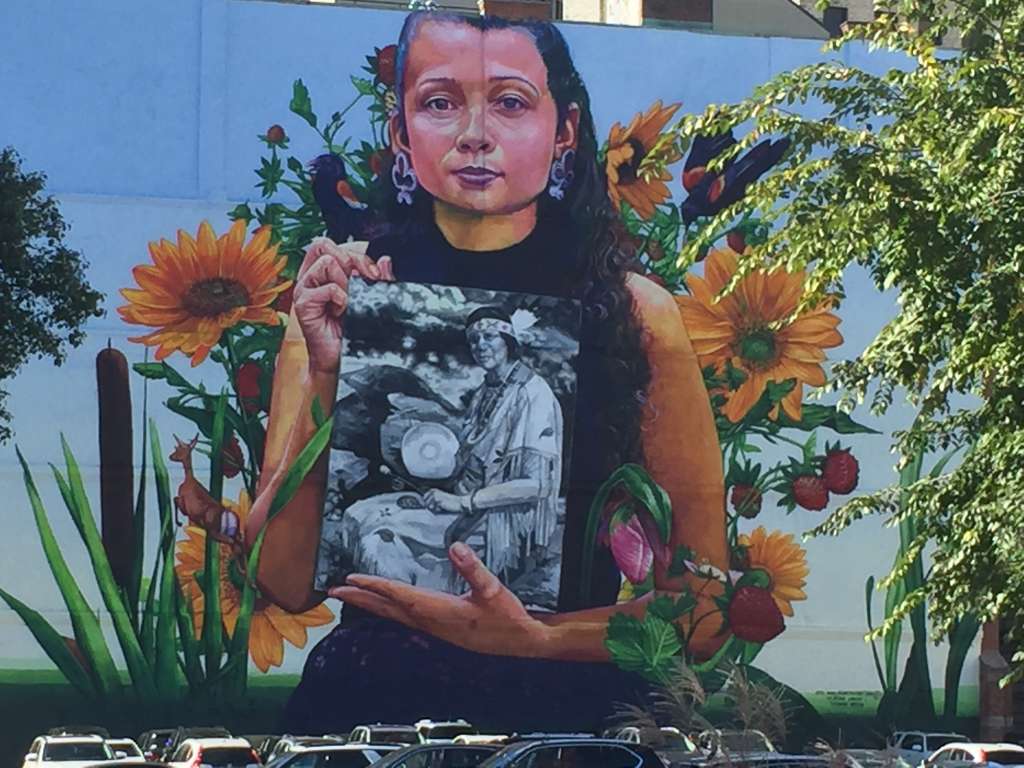 Mural (2018) depicting a member of the Narragansett Indian Tribe holding a photo of Princess Red Wing, a Narragansett/Pokanoket-Wampanoag elder who founded the Tomaquag Museum (Exeter).  Located at 32 Custom House Street.