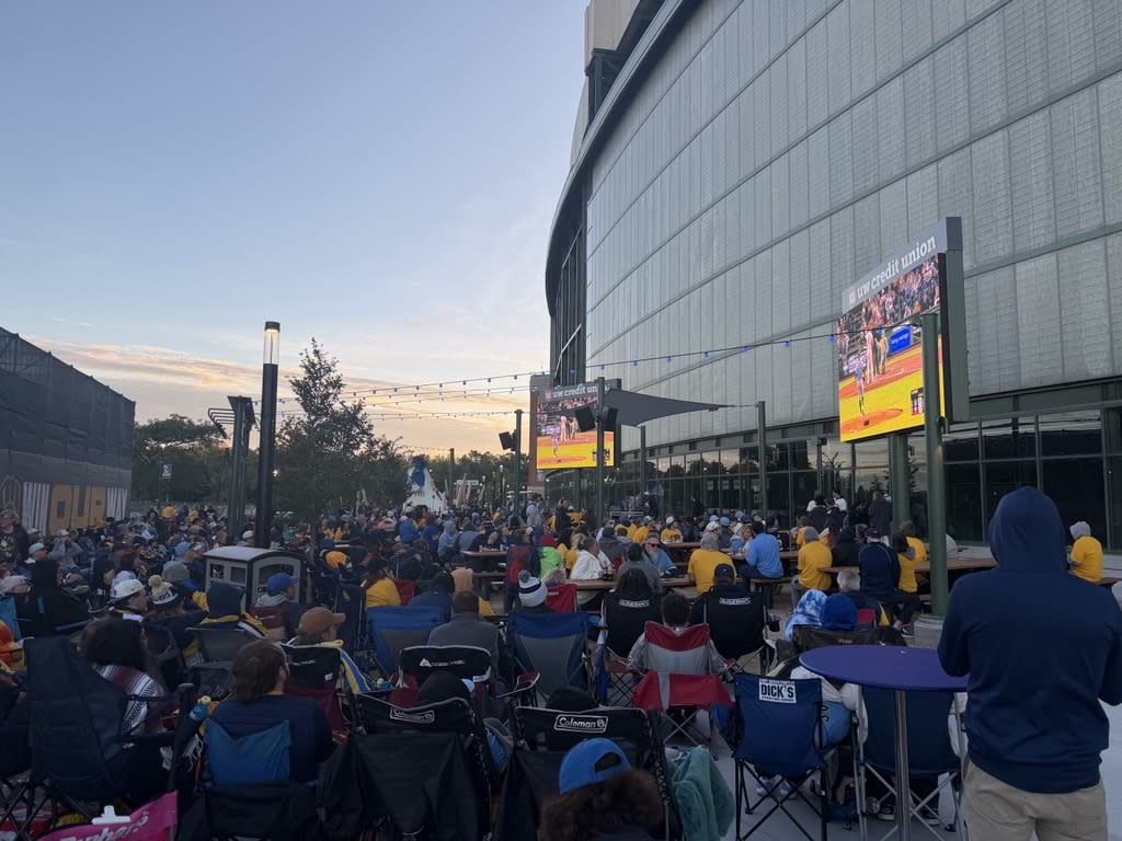 Crowd of Milwaukee Brewers fans gathered outside a stadium, watching a baseball game on large outdoor video screens at sunset.