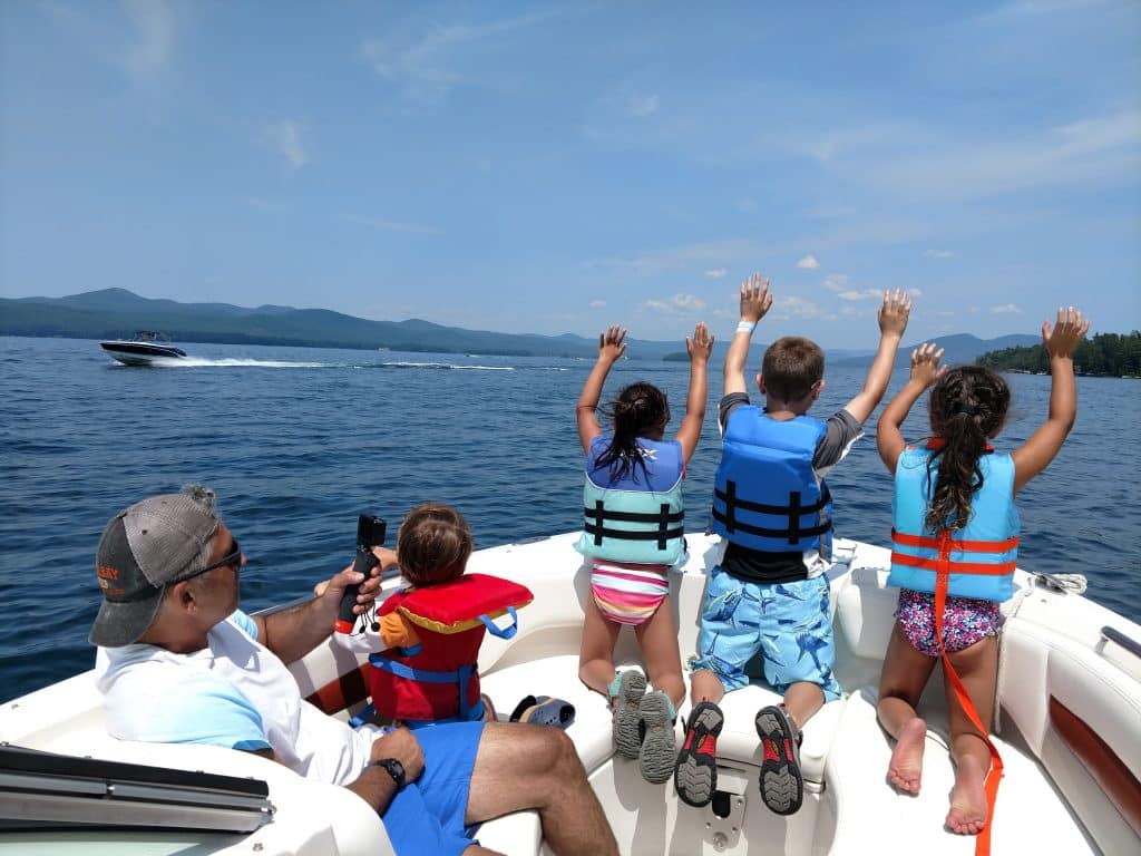 A family on a boat with kids holding their hands in the air at another boat racing by on Lake George
