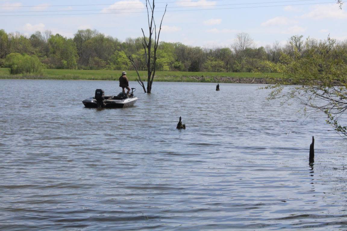 Person fishing from a small boat on a calm lake surrounded by spring trees