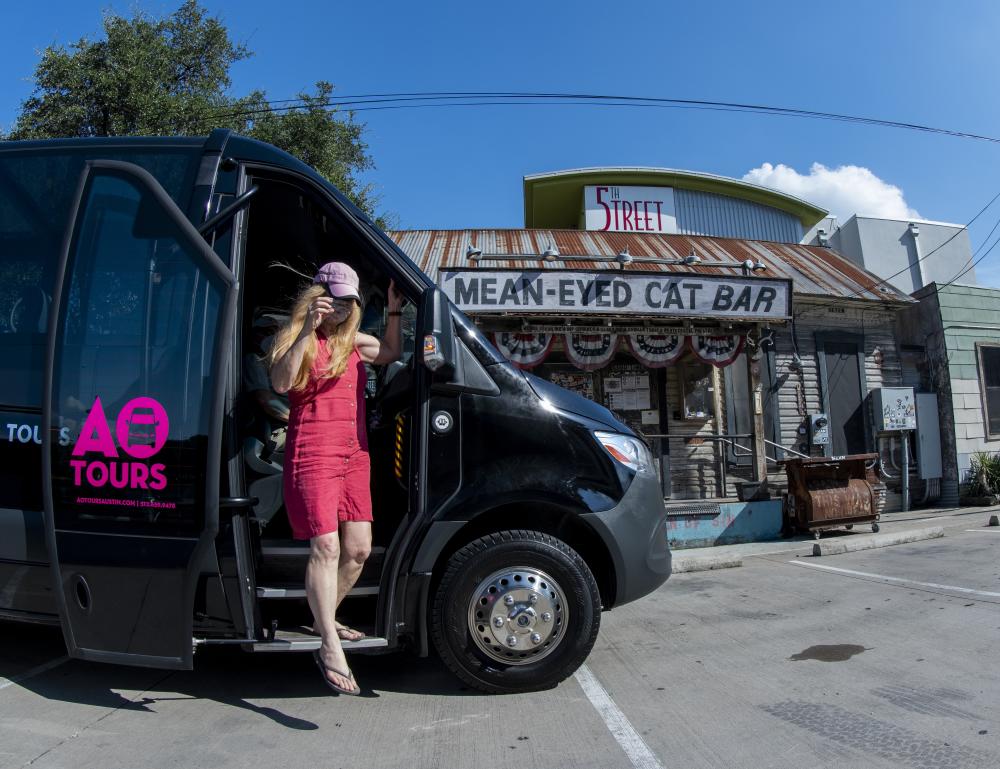 An AO Tours Van parked outside the Mean Eyed Cat bar. A woman in a pink dress is stepping down from the van