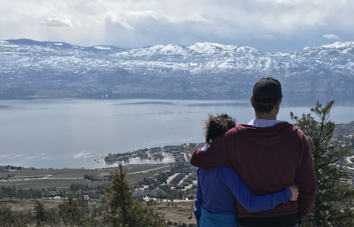 A couple holding each other stands near the top Mount Boucherie looking out over Kelowna, BC