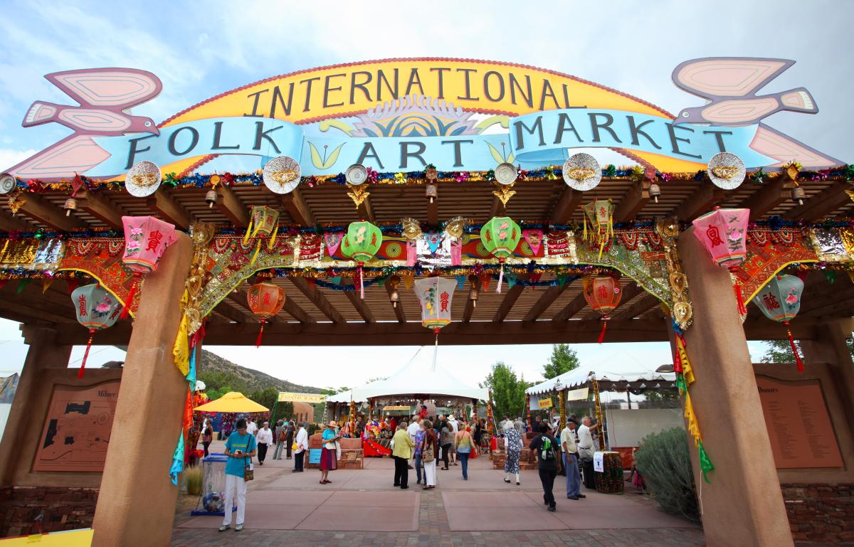 A group of people stand beneath a large archway adorned with hanging art pieces with a colorful sign that reads "International Folk Art Market"