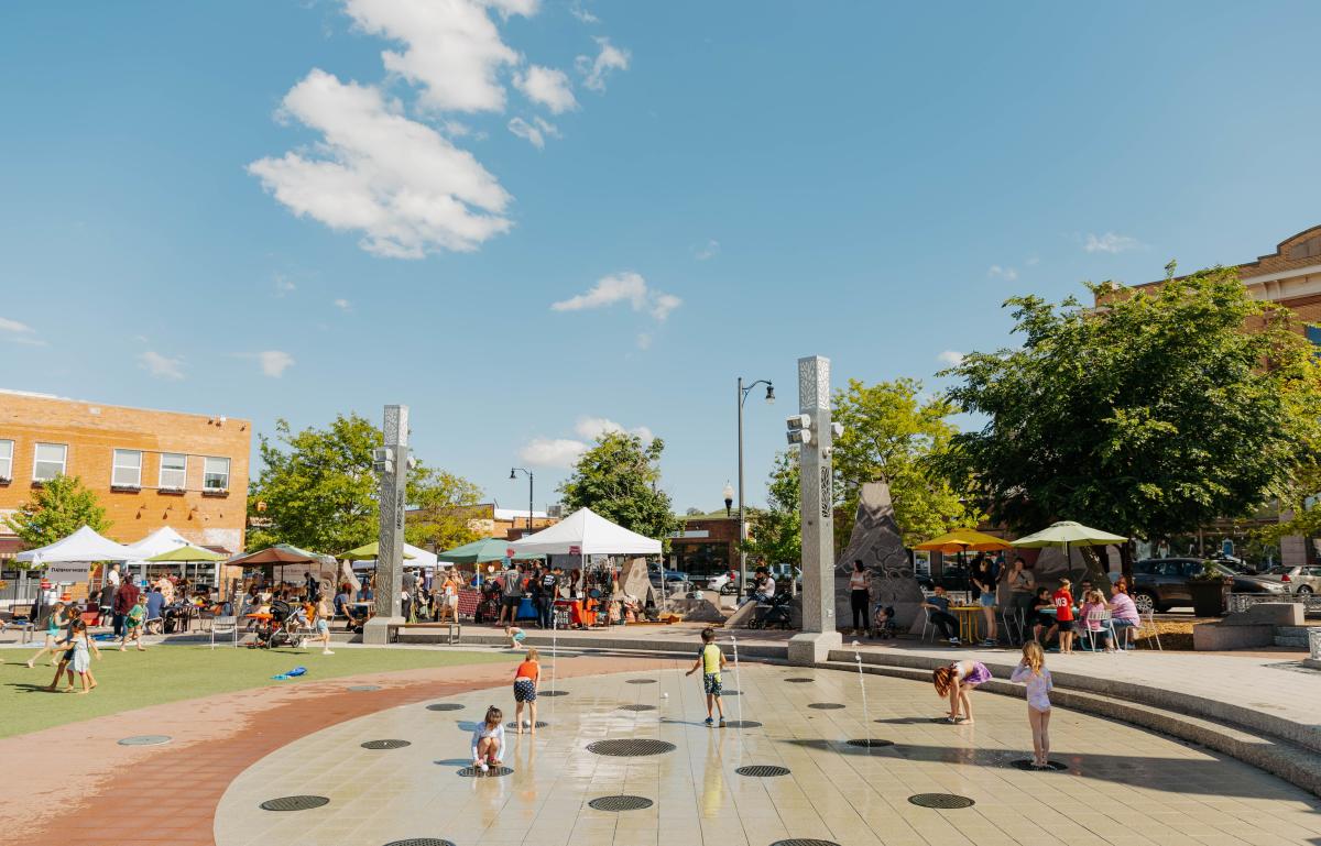 Five children play in the fountains at Main Street Square during a downtown event. White tents and many people can be seen in the background.