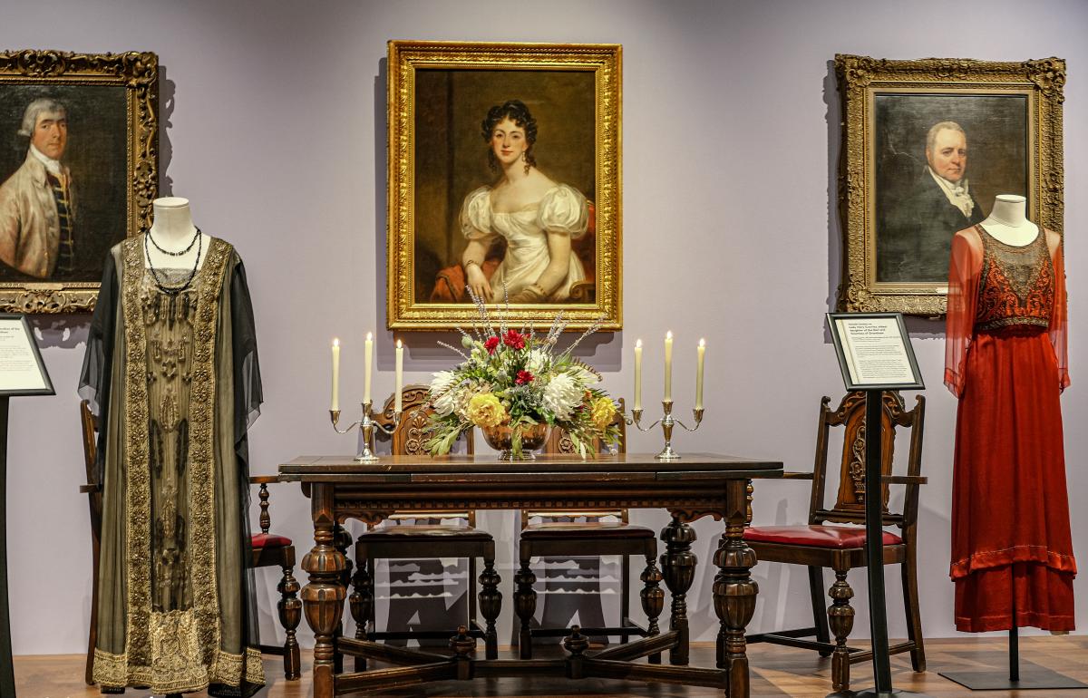 view of Muskegon Museum of Art gallery shows historic dresses flanking period dining table set with fresh flowers and candles. There are 3 portraits hanging on the wall behind the display.