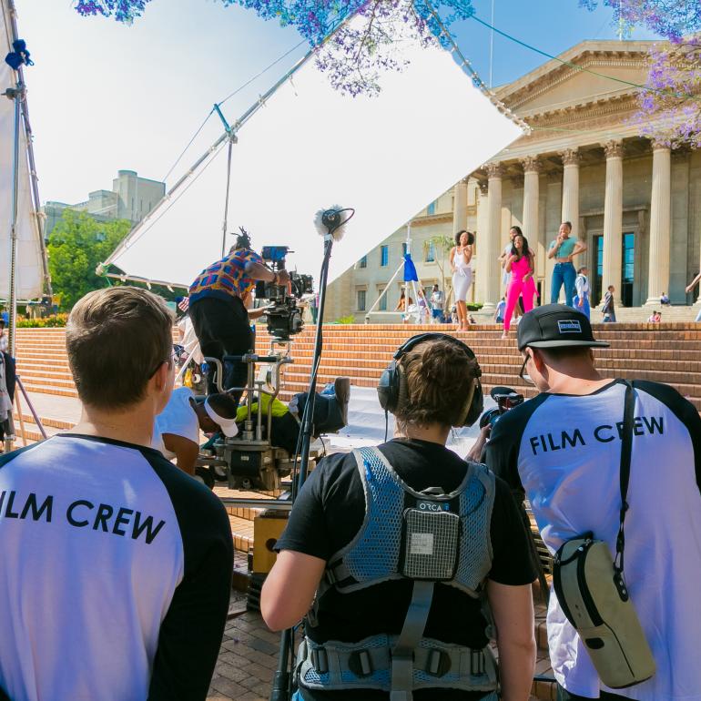 Production crew filming girls singing on the steps in front of a building with columns.