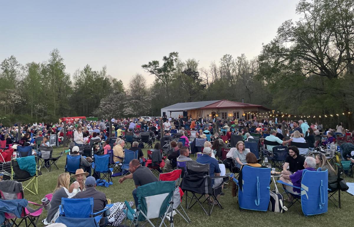 Crowd seated in lawn chairs at a Jazz’n the Vines concert at Wild Bush Farm and Vineyard, with the stage and vineyard buildings surrounded by trees at dusk.