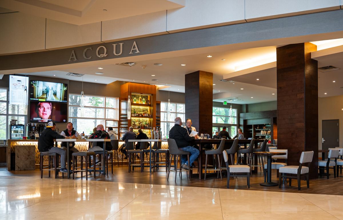 The camera faces an open room attached to lobby of The Woodlands Waterway Marriott. A dark gray sign over the room reads, in large silver text, "ACQUA." The room features a dozen or more tables of various sizes (small round tables, tall square tables, and long rectangular tables) that seat varying numbers of people. In the back left is a long, modern bar.