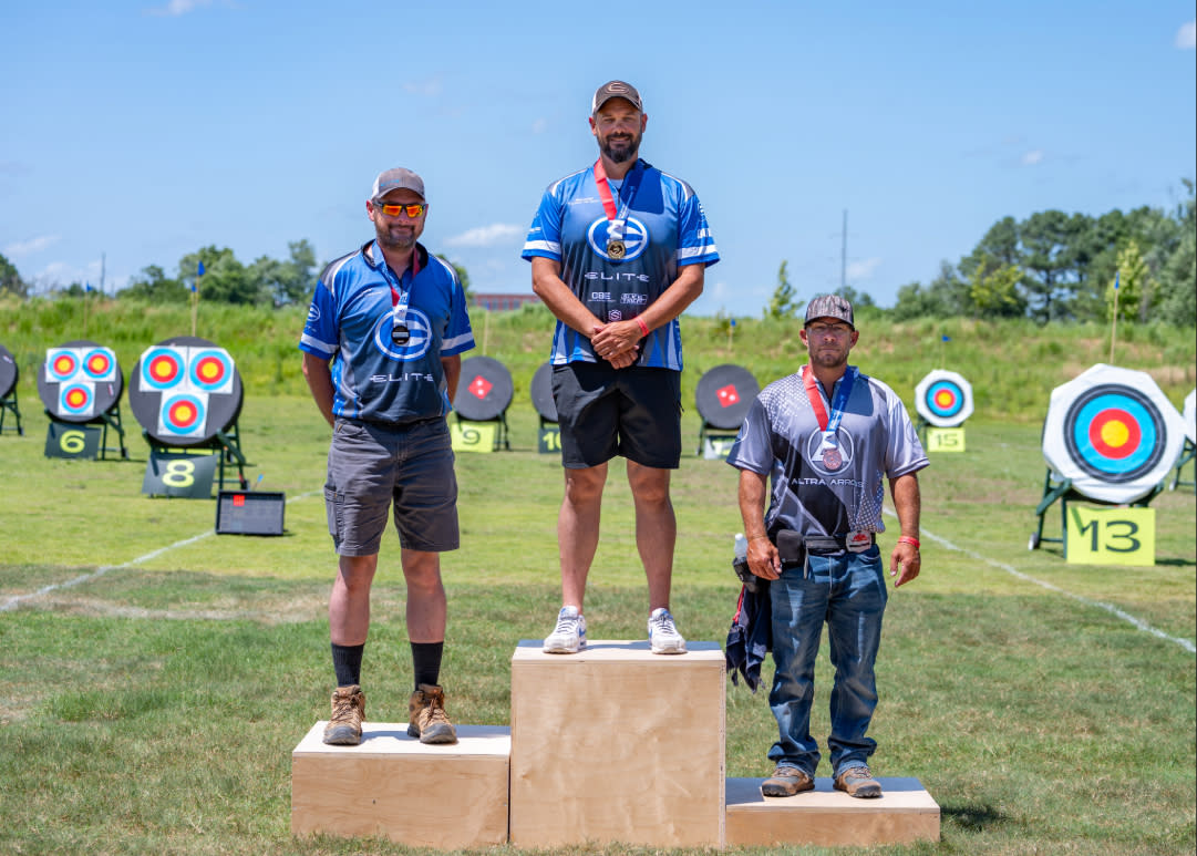 Three medal-winning archers stand on a podium at an outdoor archery range during a tournament. The first-place competitor stands on the center platform wearing a blue jersey and gold medal, while the second- and third-place finishers stand on either side with their medals. Colorful archery targets are visible in the background under a clear blue sky.