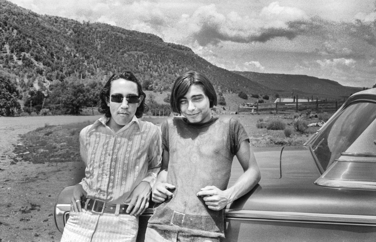 Black and white photograph of two teenage boys leaning against a parked car on a dirty roadside.