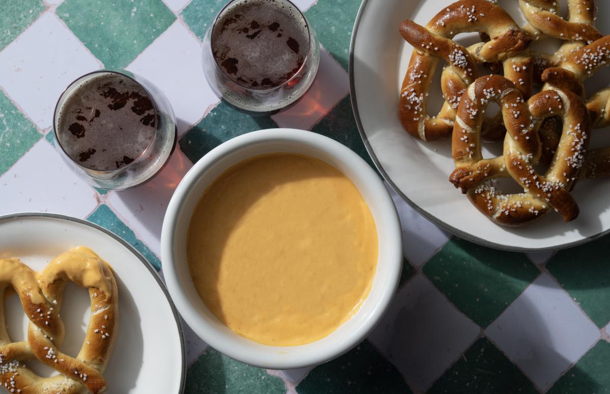 Bowl of golden beer-cheese dip on a checkered table with soft pretzels and two dark beers.