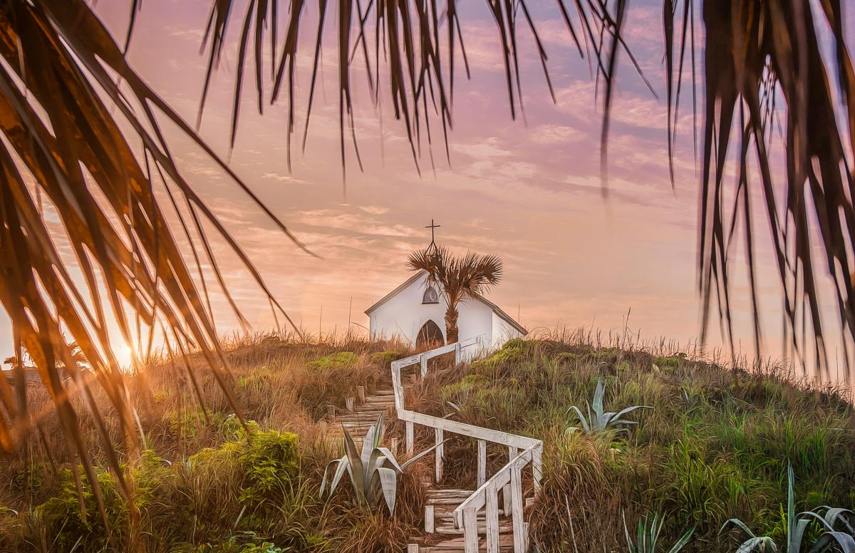 On top of a grassy hill at sunset sits a small white chapel topped with a cross. Steps with a white railing lead up to the chapel and palm tree fronds frame the photo