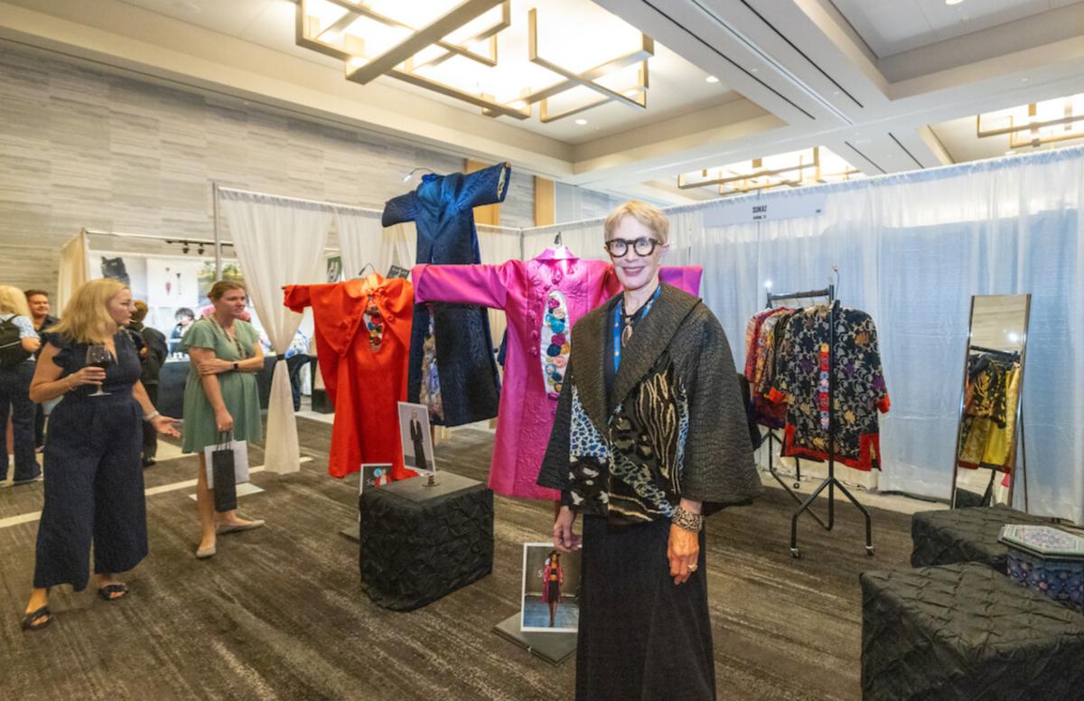 An art booth at the Texas Fine Craft Show features a prominent display of three East Asian-inspired clothing designs, one bright red, one blue, one hot pink. SuzKaz, a woman with short-cropped blonde hair, thick-rimmed glasses, and a thick animal-skin shawl, stands with her work as guests admire her work.
