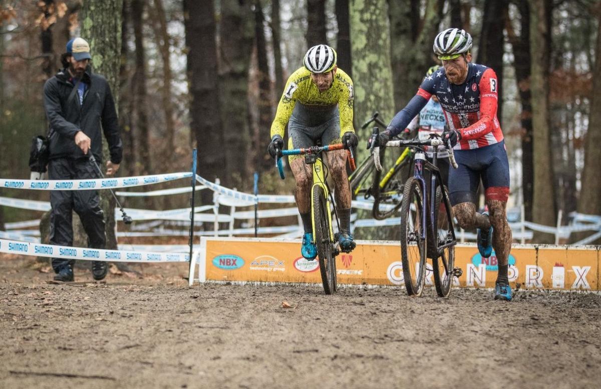 Two bikers on a dirt course, one riding his bike while the other runs alongside his own bike, with a man watching in the left background.