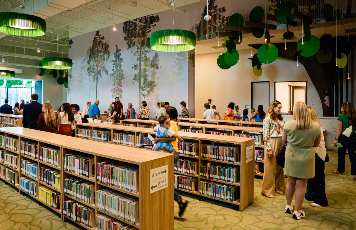 In one of the main rooms of the Kevin Brady Library are several rows of long, child-height bookshelves, several craft tables, and three padded reading nooks along the left wall. Lights hanging from the ceiling look like large lily pads. The room is full of people for the grand opening.