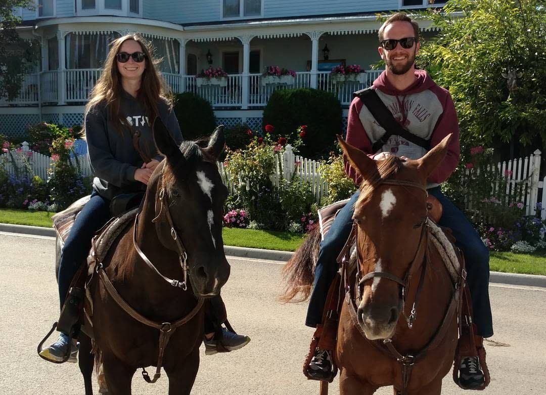 A couple smiles while riding horseback on the road out front of a Mackinac Island hotel